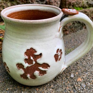 May include: Handmade ceramic mug with a light green glaze and brown poodle and paw print designs. The interior is a warm brown color. The handle has a small decorative accent. A unique, handcrafted piece for coffee or tea.