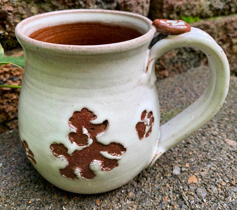 May include: Handmade ceramic mug with a light green glaze and brown poodle and paw print designs. The interior is a warm brown color. The handle has a small decorative accent. A unique, handcrafted piece for coffee or tea.