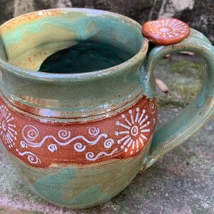 May include: A handmade ceramic mug with a green glaze and brown accents. The mug has a unique design with white swirls and a small knob on the handle.