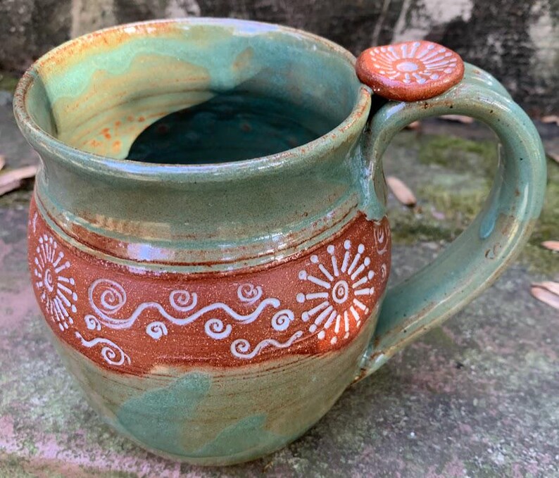 May include: A handmade ceramic mug with a green glaze and brown accents. The mug has a unique design with white swirls and a small knob on the handle.