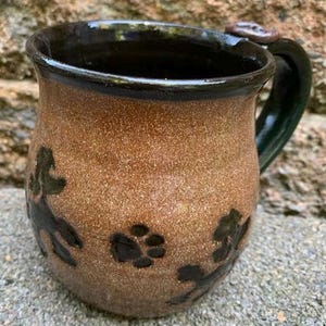 May include: A handmade ceramic mug with a brown speckled exterior and a dark green handle. The mug features black paw prints and leaf designs. The rim and interior are dark brown. This unique mug is perfect for coffee or tea.
