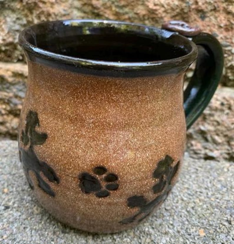 May include: A handmade ceramic mug with a brown speckled exterior and a dark green handle. The mug features black paw prints and leaf designs. The rim and interior are dark brown. This unique mug is perfect for coffee or tea.