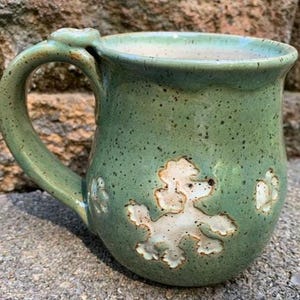 May include: A handmade ceramic mug in a speckled green glaze. The mug features a white poodle design and a curved handle. The mug is a unique piece of pottery, perfect for coffee or tea.