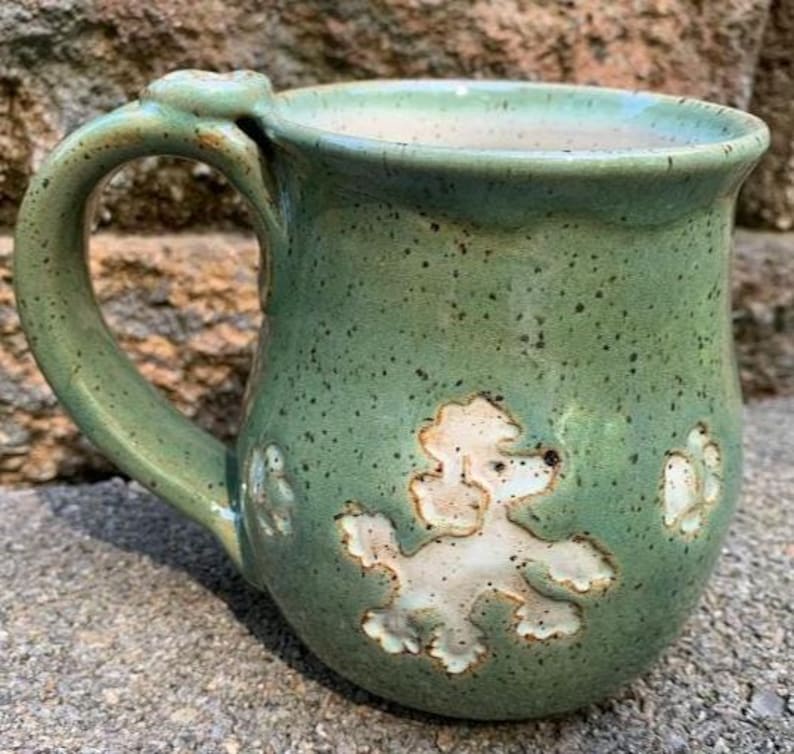 May include: A handmade ceramic mug in a speckled green glaze. The mug features a white poodle design and a curved handle. The mug is a unique piece of pottery, perfect for coffee or tea.