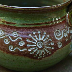May include: A green and brown ceramic mug with a white design of a stylized flower and wavy lines. The mug has a handle and a rounded shape.