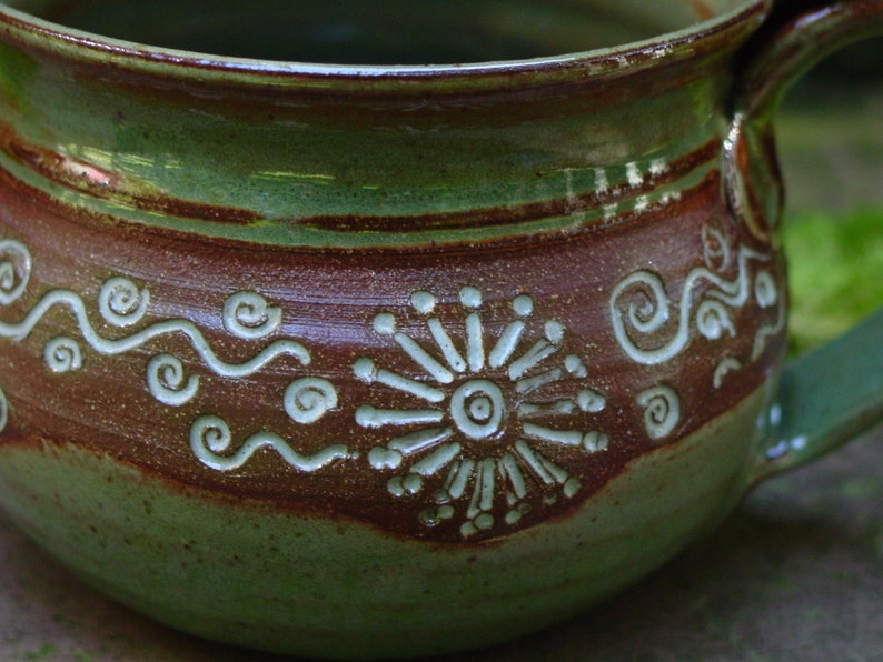 May include: A green and brown ceramic mug with a white design of a stylized flower and wavy lines. The mug has a handle and a rounded shape.