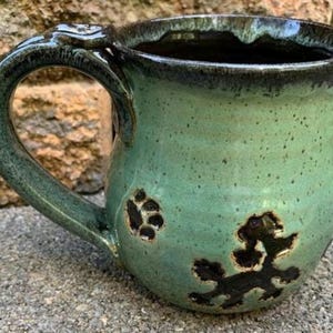 May include: Handmade ceramic mug in shades of green with a dark rim and handle. The handle features a decorative paw print design. The mug has a paw print design on the front. A unique, handcrafted piece for coffee or tea.