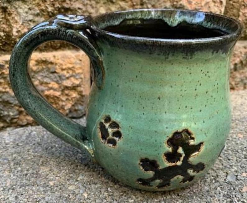 May include: Handmade ceramic mug in shades of green with a dark rim and handle. The handle features a decorative paw print design. The mug has a paw print design on the front. A unique, handcrafted piece for coffee or tea.