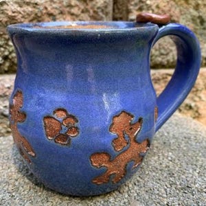 May include: A handmade blue ceramic mug with a large handle and a brown accent on the rim. The mug features brown paw print and dog-shaped designs. The mug is sitting on a stone surface.