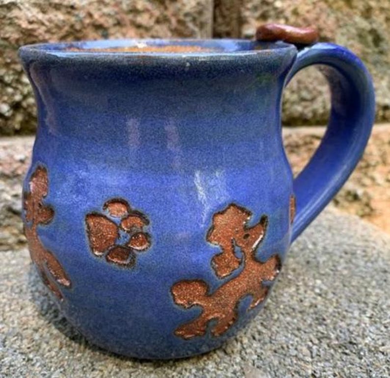 May include: A handmade blue ceramic mug with a large handle and a brown accent on the rim. The mug features brown paw print and dog-shaped designs. The mug is sitting on a stone surface.