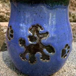 May include: A blue ceramic mug with a poodle design and paw prints. The mug has a dark blue rim and a textured surface. The poodle and paw prints are dark brown.