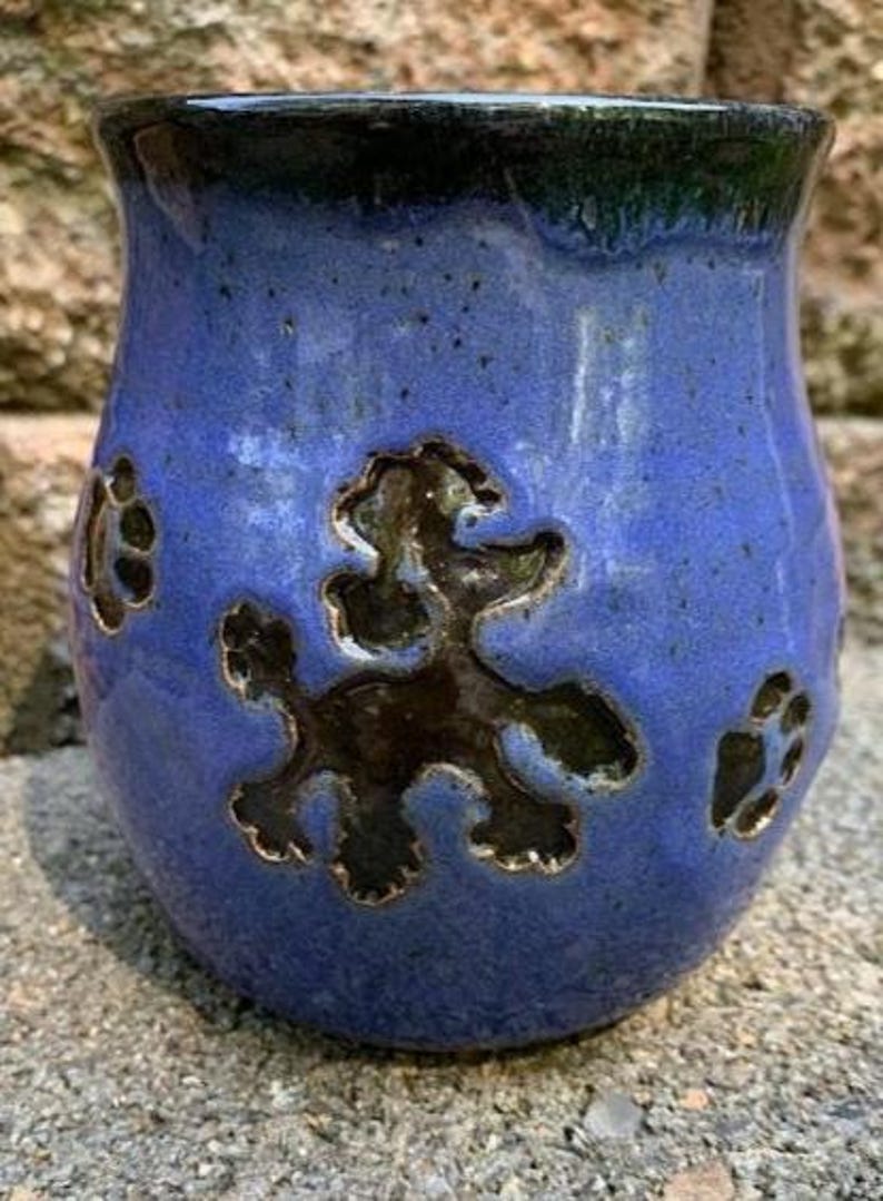 May include: A blue ceramic mug with a poodle design and paw prints. The mug has a dark blue rim and a textured surface. The poodle and paw prints are dark brown.