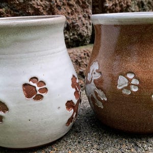 May include: Two ceramic mugs with handles. One is off-white with brown paw prints and poodle designs. The other is brown with white paw prints and poodle designs. Both mugs have a light brown rim and handle accents.