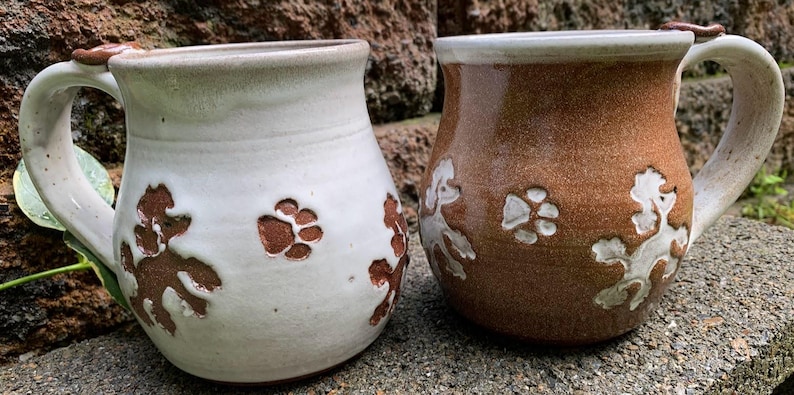 May include: Two ceramic mugs with handles. One is off-white with brown paw prints and poodle designs. The other is brown with white paw prints and poodle designs. Both mugs have a light brown rim and handle accents.