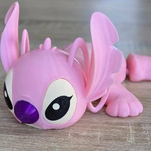May include: A pink 3D-printed figurine of a cartoon character with large ears, large eyes, and a purple nose. The figurine is lying down, with its arms and legs extended. The character's eyes are black and white.