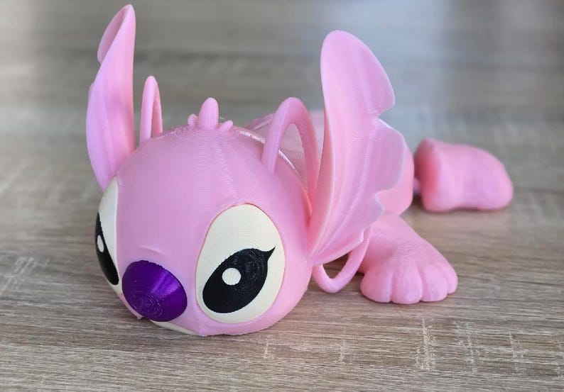 May include: A pink 3D-printed figurine of a cartoon character with large ears, large eyes, and a purple nose. The figurine is lying down, with its arms and legs extended. The character's eyes are black and white.