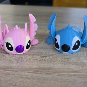May include: Two 3D-printed figurines of the character Stitch, one pink and one blue, are displayed on a wooden surface. The figurines have large ears, black eyes, and a purple nose. The blue figurine is on the right, and the pink figurine is on the left.