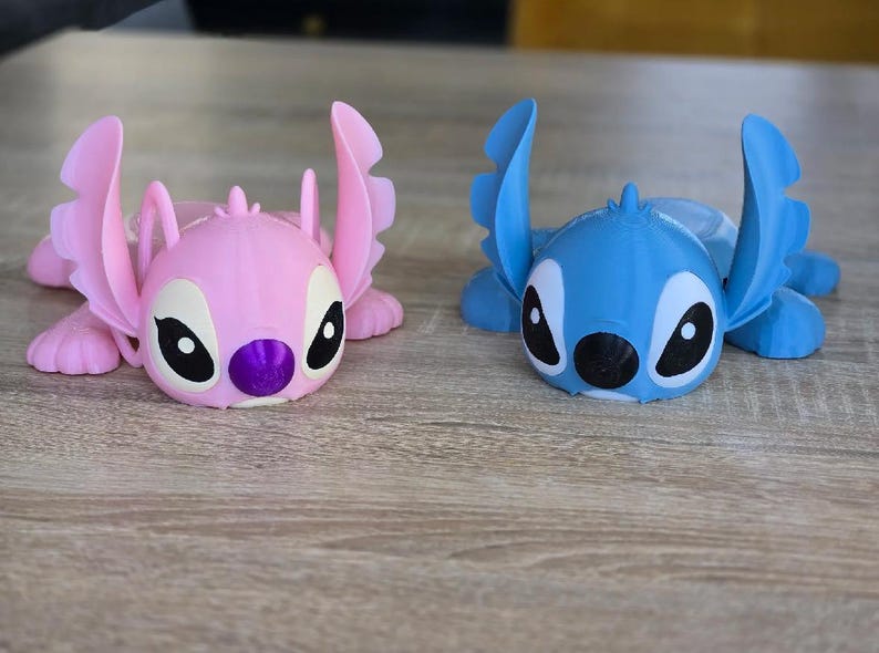 May include: Two 3D-printed figurines of the character Stitch, one pink and one blue, are displayed on a wooden surface. The figurines have large ears, black eyes, and a purple nose. The blue figurine is on the right, and the pink figurine is on the left.