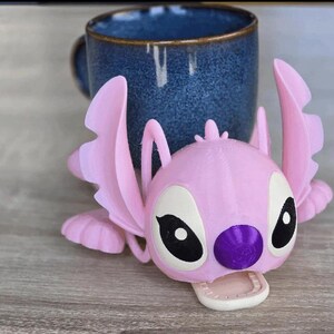 May include: A pink 3D-printed Angel figure, with large ears and wings, rests in front of a blue ceramic mug. The figure has black and white eyes, a purple nose, and a white mouth. The mug is dark blue with a textured surface.