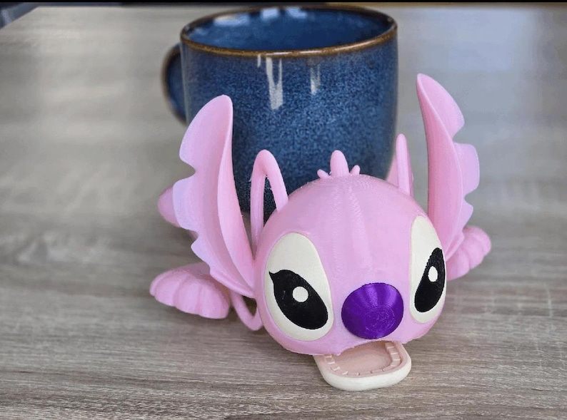 May include: A pink 3D-printed Angel figure, with large ears and wings, rests in front of a blue ceramic mug. The figure has black and white eyes, a purple nose, and a white mouth. The mug is dark blue with a textured surface.