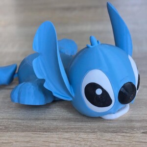 May include: A light blue figurine of the character Stitch from the animated movie Lilo & Stitch. The figurine is lying down with its large ears and distinctive features clearly visible. The eyes are white with black pupils, and the mouth is white.