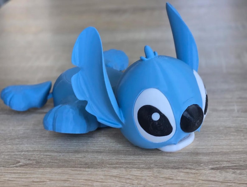May include: A light blue figurine of the character Stitch from the animated movie Lilo & Stitch. The figurine is lying down with its large ears and distinctive features clearly visible. The eyes are white with black pupils, and the mouth is white.