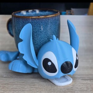 May include: A blue ceramic mug with a textured finish, paired with a light blue 3D-printed Stitch character. The character is lying down, with large black and white eyes and a white mouth. The mug is dark blue with a brown rim.