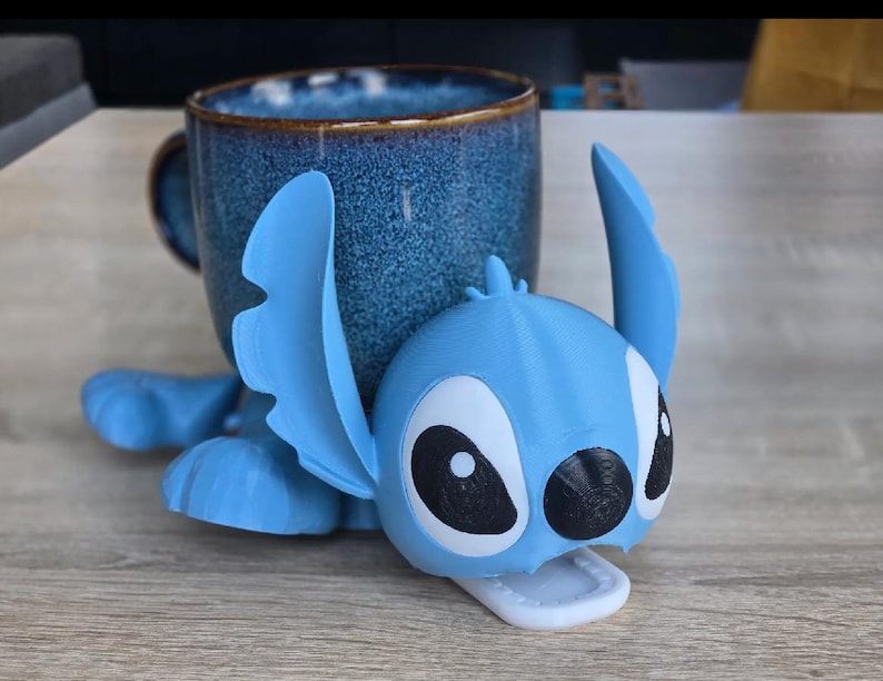 May include: A blue ceramic mug with a textured finish, paired with a light blue 3D-printed Stitch character. The character is lying down, with large black and white eyes and a white mouth. The mug is dark blue with a brown rim.