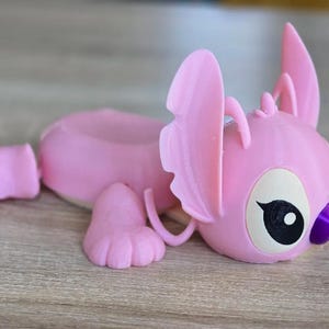 May include: A pink, 3D-printed figurine of a cartoon character, lying down. The figurine has large ears, a round head, and a small purple nose. The eyes are black with white highlights. The figurine is on a wooden surface.