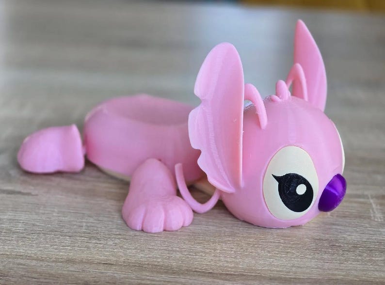 May include: A pink, 3D-printed figurine of a cartoon character, lying down. The figurine has large ears, a round head, and a small purple nose. The eyes are black with white highlights. The figurine is on a wooden surface.