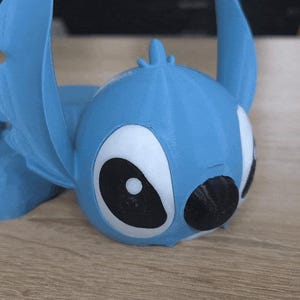 May include: A light blue 3D-printed figurine of a cartoon character with large black and white eyes and large ears. The figurine is lying down on a wooden surface. The character is a popular animated character.