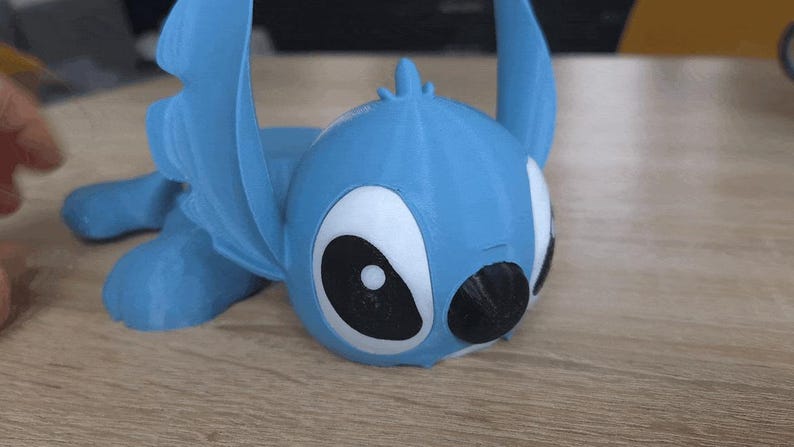 May include: A light blue 3D-printed figurine of a cartoon character with large black and white eyes and large ears. The figurine is lying down on a wooden surface. The character is a popular animated character.