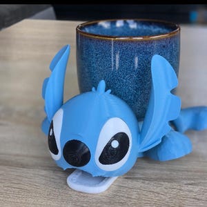 May include: A light blue 3D-printed character with large ears and black eyes, resembling Stitch from Lilo & Stitch. The character is positioned in front of a blue ceramic mug with a brown rim. The mug has a textured surface.
