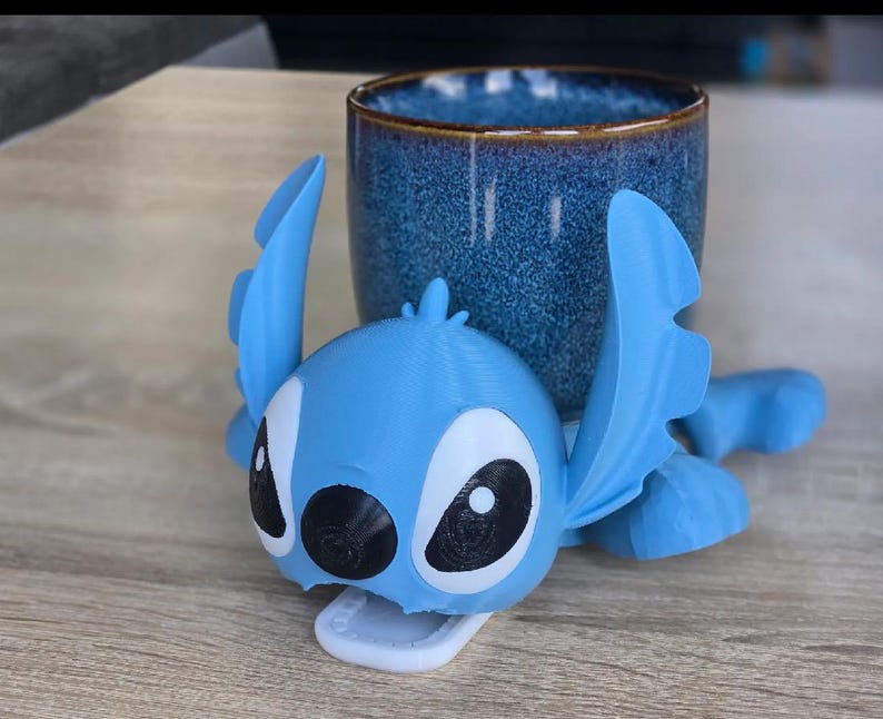 May include: A light blue 3D-printed character with large ears and black eyes, resembling Stitch from Lilo & Stitch. The character is positioned in front of a blue ceramic mug with a brown rim. The mug has a textured surface.