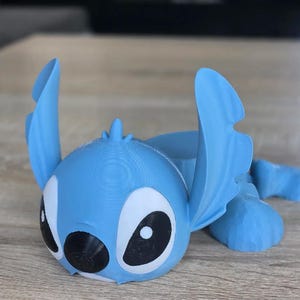 May include: A light blue 3D-printed figurine of the character Stitch from the Disney movie Lilo & Stitch. The figurine has large black and white eyes, large ears, and is lying down. The figurine is made of plastic.