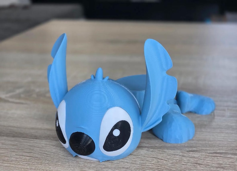 May include: A light blue 3D-printed figurine of the character Stitch from the Disney movie Lilo & Stitch. The figurine has large black and white eyes, large ears, and is lying down. The figurine is made of plastic.