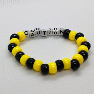 Puede incluir: Una pulsera de cuentas amarillas y negras, con la palabra "CAUTION" escrita con cuentas de letras blancas. Las cuentas están ensartadas para formar una forma circular. La pulsera es un accesorio de moda, probablemente para fines de novedad.