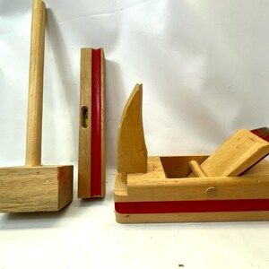 May include: A collection of wooden toy tools, including a mallet, a level, and a plane. The tools are made of light-colored wood with red accents. The mallet has a rectangular head, and the level has a bubble indicator.