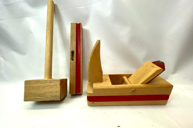 May include: A collection of wooden toy tools, including a mallet, a level, and a plane. The tools are made of light-colored wood with red accents. The mallet has a rectangular head, and the level has a bubble indicator.