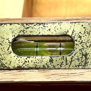 May include: Close-up of a vintage level tool. The level has a wooden base with a distressed, speckled, gold-colored metal top. A clear vial with a green liquid and black lines indicates the level. The tool is used for construction and carpentry.
