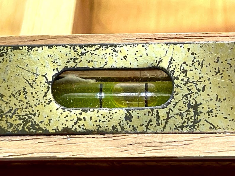 May include: Close-up of a vintage level tool. The level has a wooden base with a distressed, speckled, gold-colored metal top. A clear vial with a green liquid and black lines indicates the level. The tool is used for construction and carpentry.