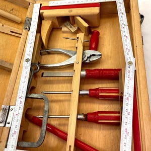 May include: A wooden toolbox filled with various hand tools. The tools include pliers, screwdrivers with red handles, and a metal clamp. A folding ruler is visible along the side. The box is made of light-colored wood.