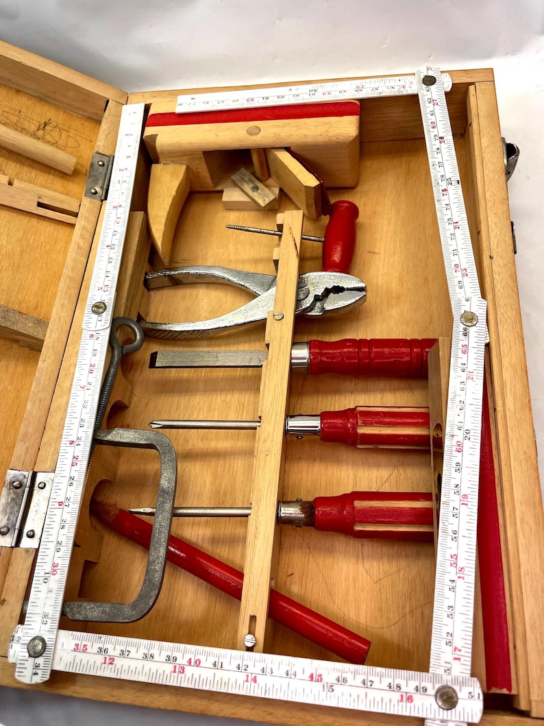 May include: A wooden toolbox filled with various hand tools. The tools include pliers, screwdrivers with red handles, and a metal clamp. A folding ruler is visible along the side. The box is made of light-colored wood.