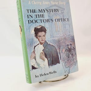May include: A vintage book titled "The Mystery in the Doctor's Office" by Helen Wells. The cover features a nurse in a white uniform and a blue coat, with a background illustration of a building and people. The book is propped up on a metal stand.