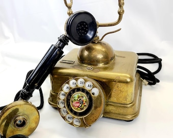 May include: A vintage brass rotary dial phone with a black handset and a white dial with black lettering. The phone has a decorative design with a floral motif on the dial.