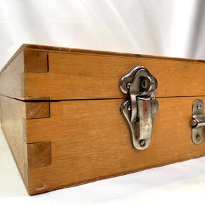 May include: A wooden box with dovetail joints, a metal clasp, and a handle. The box is made of light-colored wood and has a rectangular shape. The metal hardware is silver-colored.