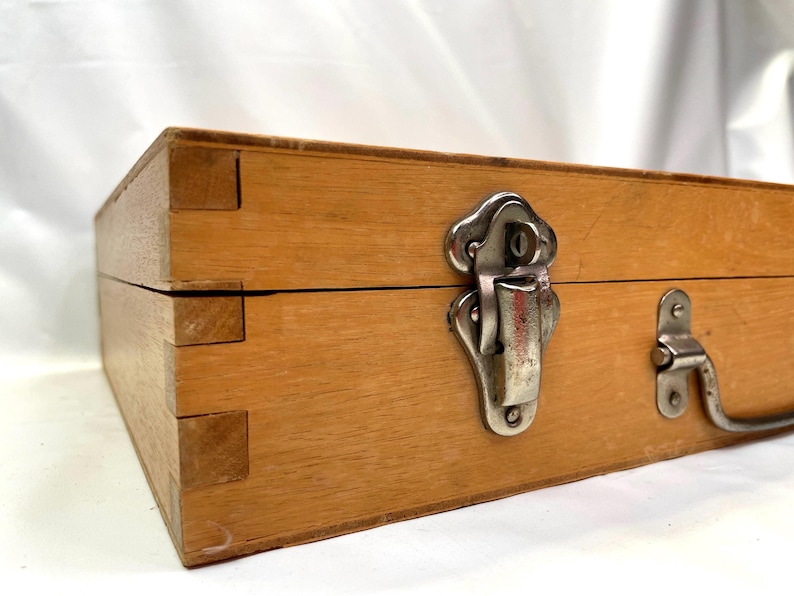 May include: A wooden box with dovetail joints, a metal clasp, and a handle. The box is made of light-colored wood and has a rectangular shape. The metal hardware is silver-colored.