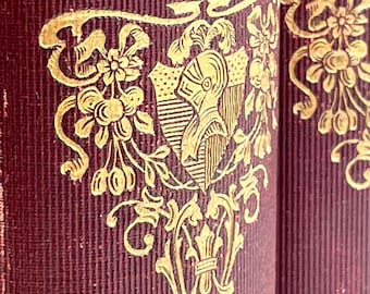 May include: A close-up of the spine of a red book with gold lettering that reads "VOL. II". The book has a decorative gold design with a shield and floral motifs.