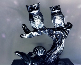 May include: A silver-colored sculpture of two owls perched on a tree branch. The owls have detailed feathers and large eyes. A mushroom and leaves are at the base of the tree. The sculpture is mounted on a black base.
