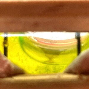 May include: A close-up view of a level tool, showing a yellow liquid bubble within a curved glass vial. The vial is set within a wooden frame, with the bubble centered, indicating a level surface.